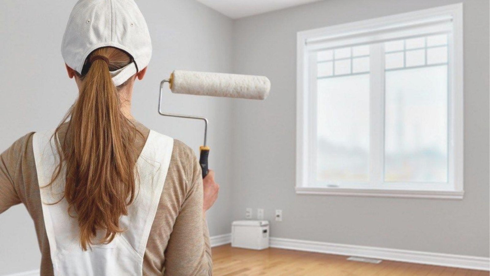 Woman in cap and overalls holding a paint roller, facing a light gray painted room.