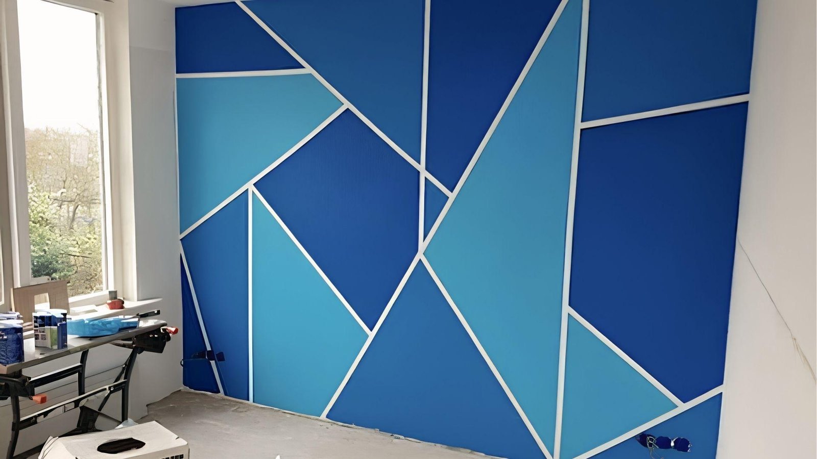 Bright blue geometric tape accent wall with white lines and paint supplies by window in progress