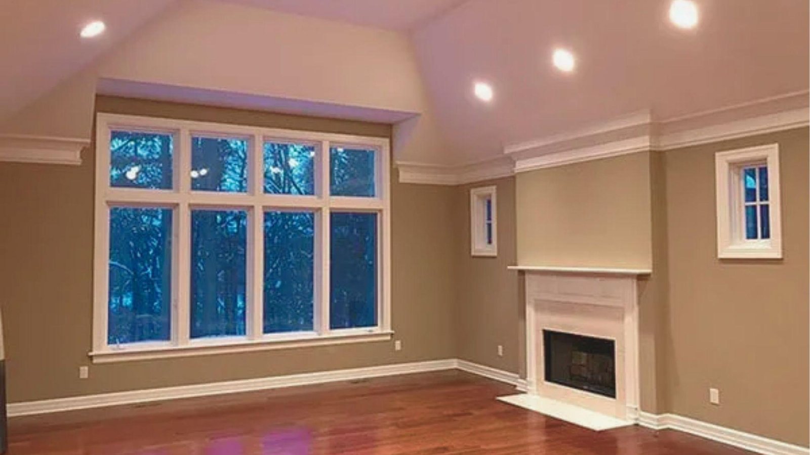 Empty living room with taupe painted walls, white trim, large windows, and a fireplace.