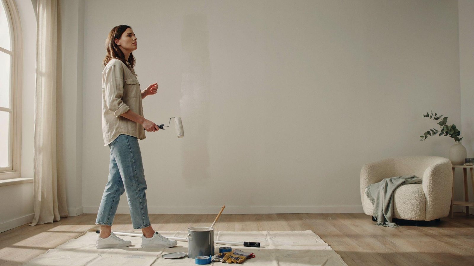 Homeowner inside a modern room holding a paint roller and considering an exterior paint can before painting an interior wall.
