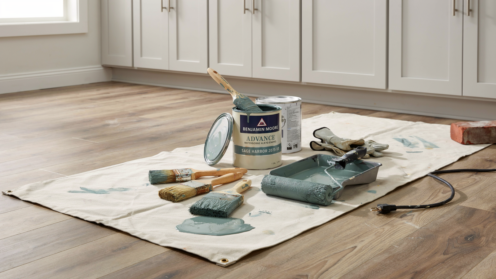 Kitchen cabinet painting tools including brushes rollers and paint cans arranged neatly on the floor near cabinets in clean indoor space.