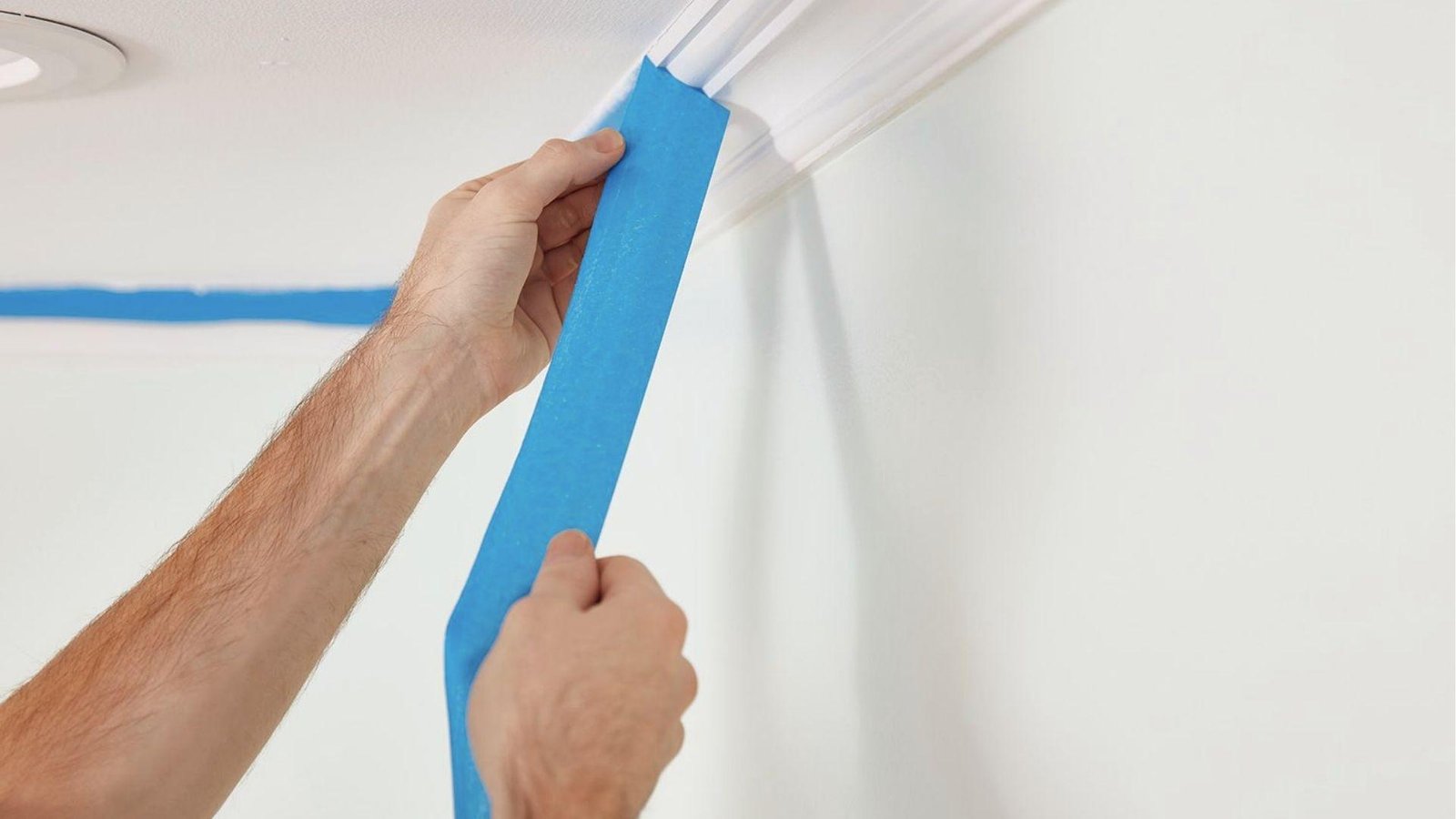Hands applying blue painter's tape along ceiling trim to prep wall for clean paint pattern edges