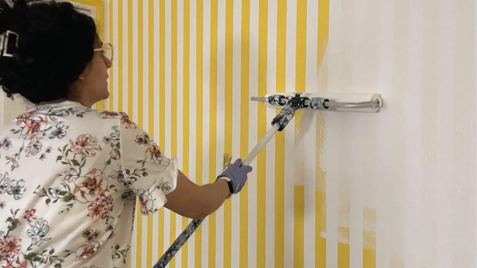 Woman painting over yellow stripe tape pattern with white roller to create crisp vertical lines