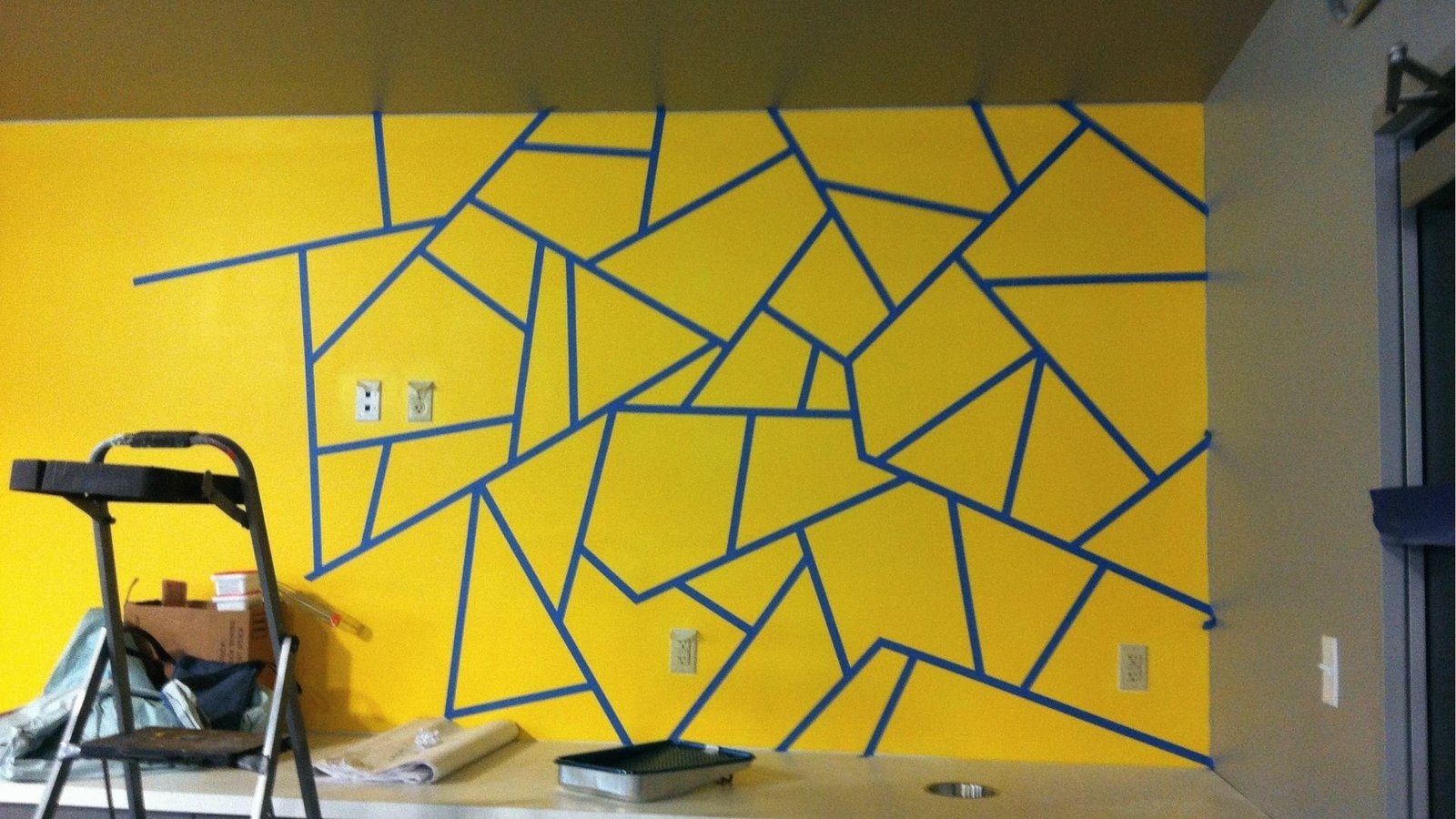 Yellow accent wall with random blue tape pattern and ladder ready for painting geometric shapes