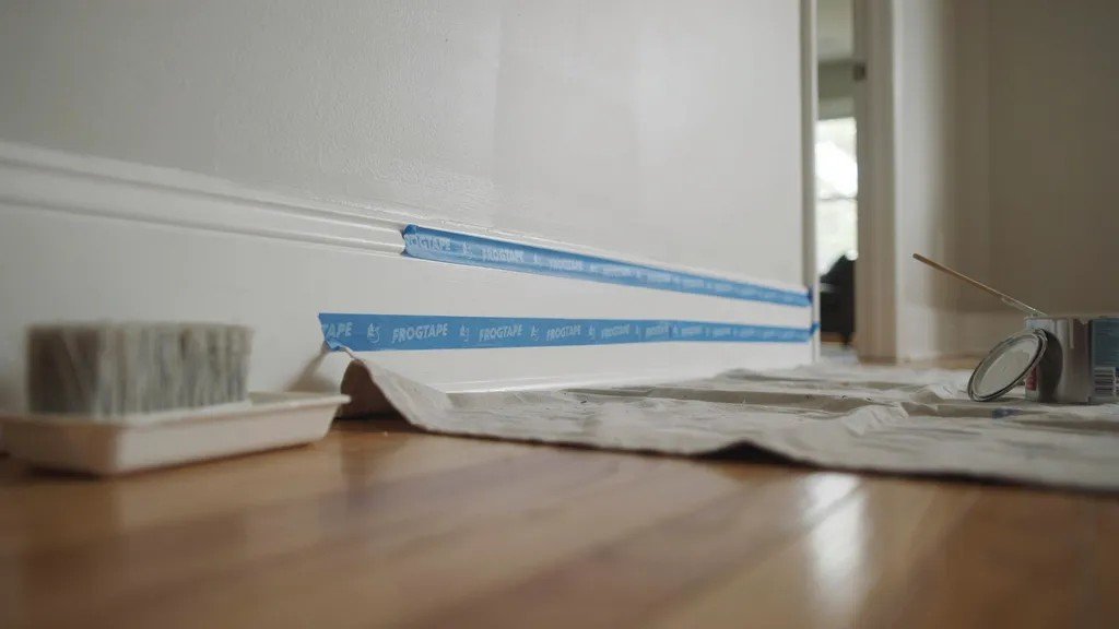 Baseboards taped along edges with floor protected using drop cloth ensuring clean paint lines in indoor setup.