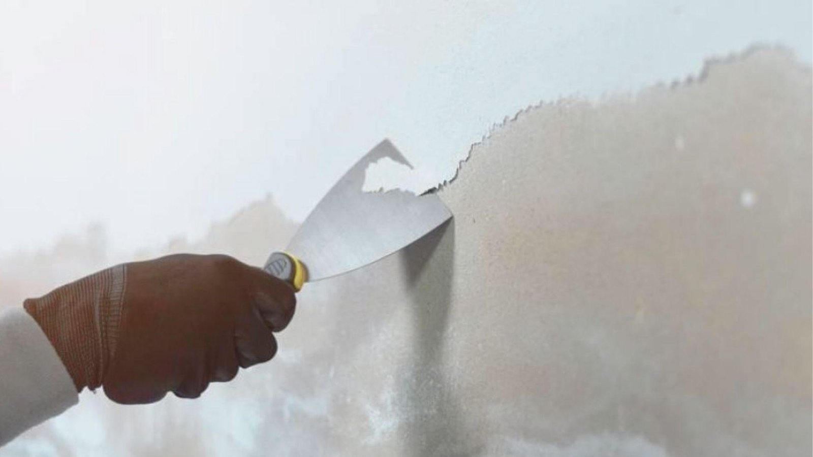 Gloved hand using a putty knife to scrape peeling white paint off an interior wall.