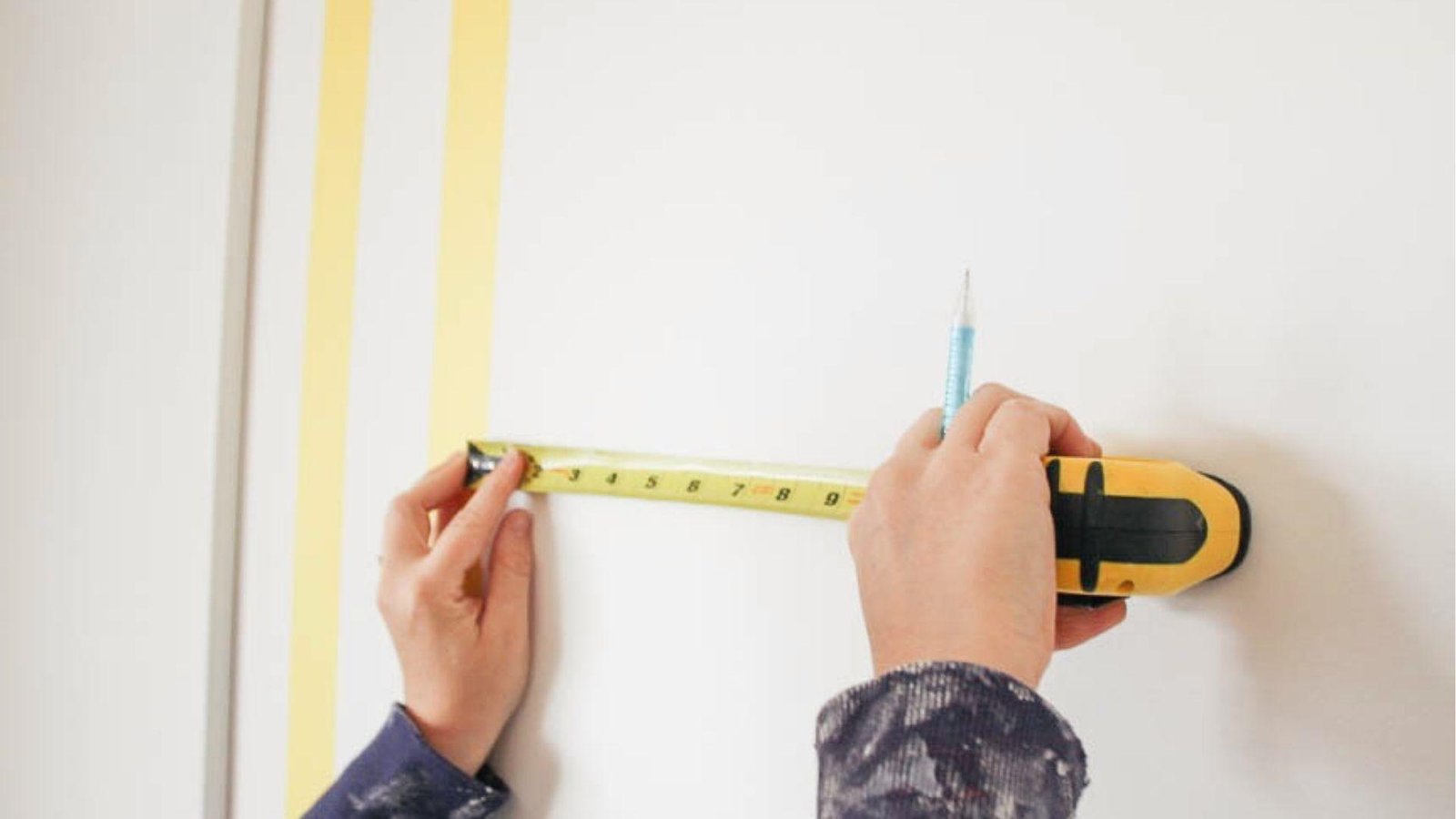Hands using measuring tape and pencil to mark even spacing for vertical tape stripes on wall