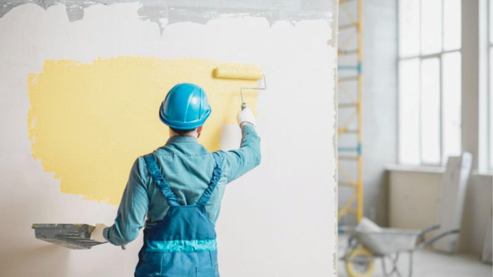 Painter in blue overalls and hard hat rolling yellow interior paint onto a white wall.